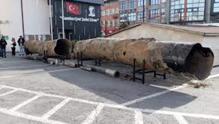 Büyük, paslanmış metal boru, kaldırım kenarında metal ayaklar üzerinde uzanıyor; arkada camlı bina bulunuyor.