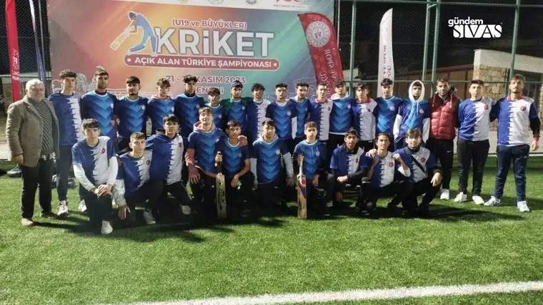 Suni çim sahada mavi formalar giyen bir grup sporcu, kriket şampiyonası pankartının önünde poz veriyor.