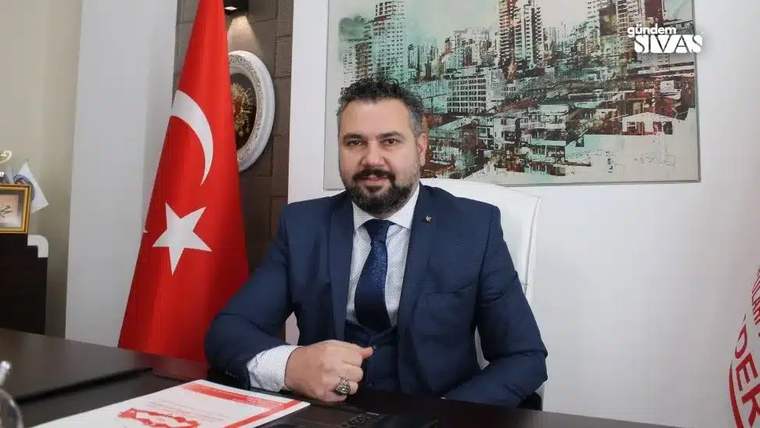 Masa başında takım elbiseli bir kişi oturuyor; arkasında Türk bayrağı ve duvarda şehir silüeti resmi bulunuyor.