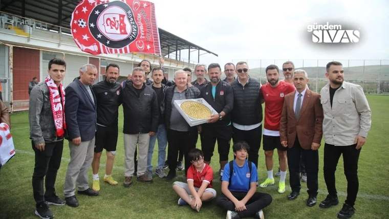 Çim sahada, üzerinde Sivasspor atkısı ve bayrağı bulunan kalabalık bir grup, tepside tatlı tutuyor.