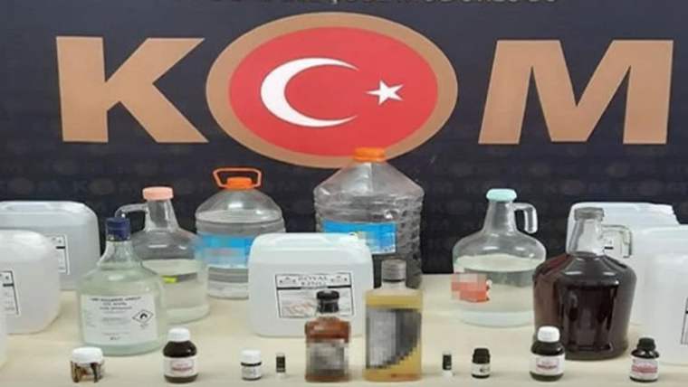 Masa üzerinde çeşitli şişe ve bidonlar, farklı renklerde sıvılar ve arkada Türk bayrağı simgesi görülüyor.
