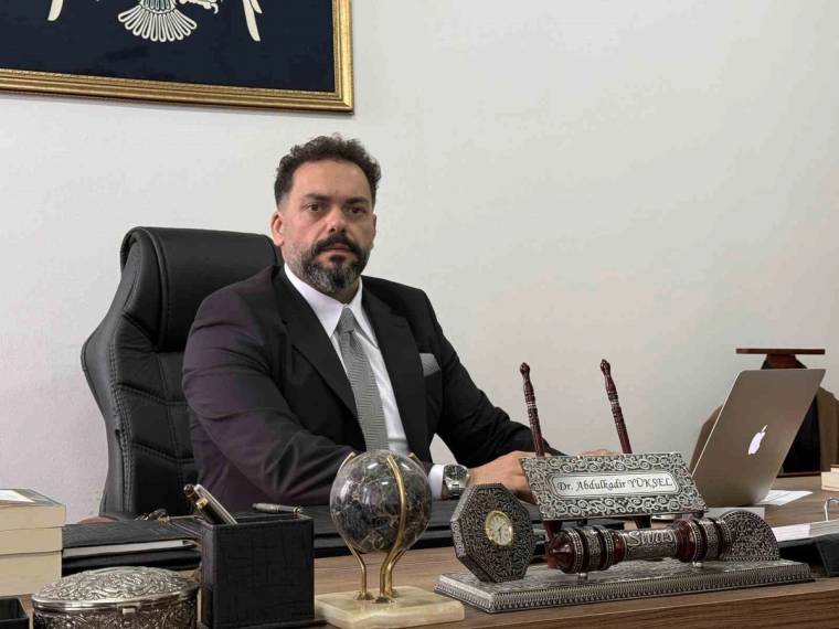 Masa başında oturan takım elbiseli bir kişi; önünde dizüstü bilgisayar, kitaplar, küre ve dekoratif eşyalar bulunuyor.