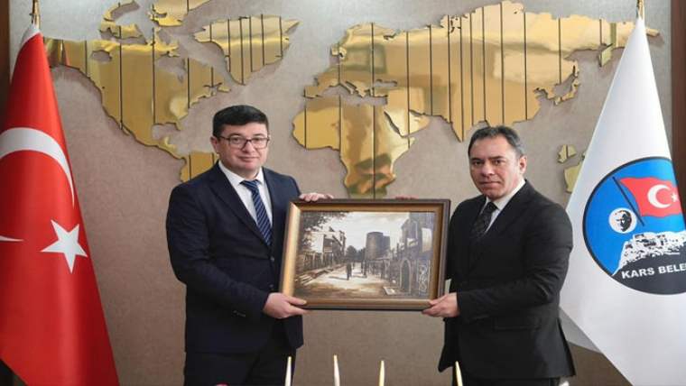 Duvarında dünya haritası bulunan salonda iki kişi, şehir manzarası içeren çerçeveyi birlikte tutuyor.