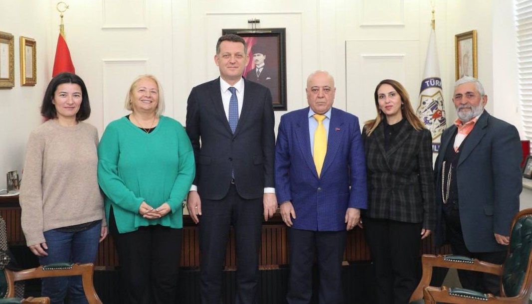 Bir odada yan yana duran altı kişi, arkalarında Türk bayrağı, duvar portresi ve kurum amblemi görülüyor.