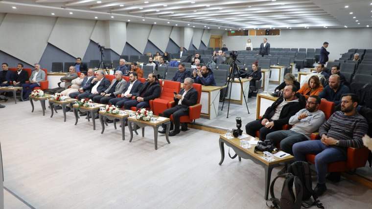 Konferans salonunda ön sırada oturan kişiler, arkalarında kameralar ve izleyicilerle birlikte görülüyor.