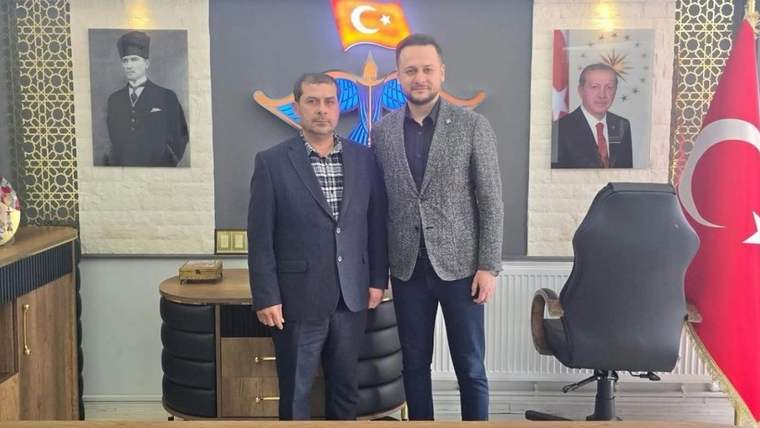 Makam odasında iki erkek, Türk bayrağı ve parti ambleminin bulunduğu duvar önünde yan yana duruyor.