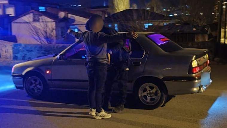Gece bir caddede, iki kişinin yanında mavi ışıklarla aydınlatılan gri bir sedan otomobil duruyor.