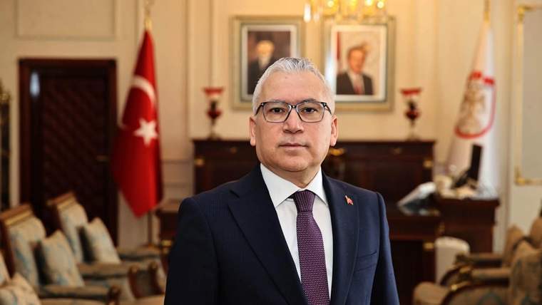 Takım elbiseli bir kişi, arkasında Türk bayrağı ve tablolar bulunan bir salonda ayakta duruyor.