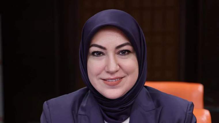 Başında başörtüsü olan bir kadın, gülümseyerek kameraya bakıyor.