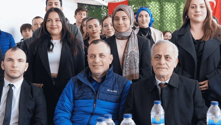 Kapalı bir alanda, masada oturan üç kişi ile arkalarında ayakta duran kalabalık bir grup görülüyor.