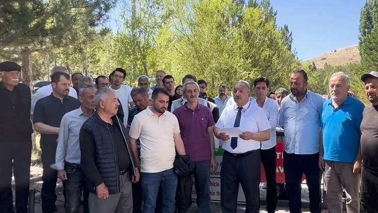 Bir grup erkek, açık havada bir araya gelerek bir kişiyi dinliyor. Arka planda ağaçlar ve dağlar görünmekte.