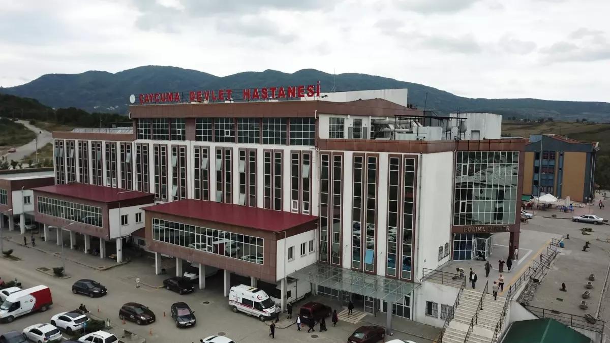 Beyaz ve kırmızı renkli bir hastane binası, önünde araçlar ve insanlar var.