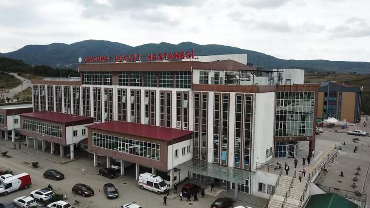 Beyaz ve kırmızı renkli bir hastane binası, önünde araçlar ve insanlar var.