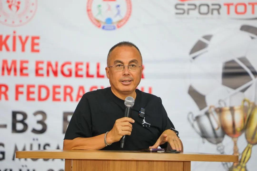 Mikrofonun önünde konuşan bir kişi, arka planda futbol temalı afişler var.