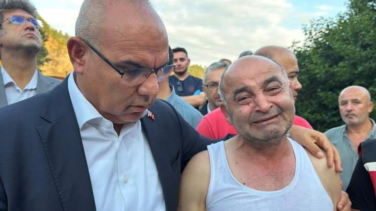 Kalabalık arasında bir adam gözyaşlarıyla dururken, yanındaki takım elbiseli kişi elini omzuna koyuyor.