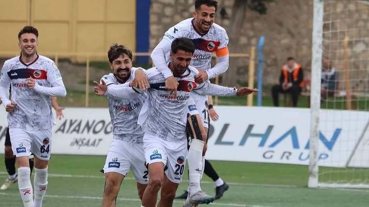 Beyaz formalar giyen futbolcular, saha içinde birbirlerine sarılarak sevinç gösterisi yapıyor.