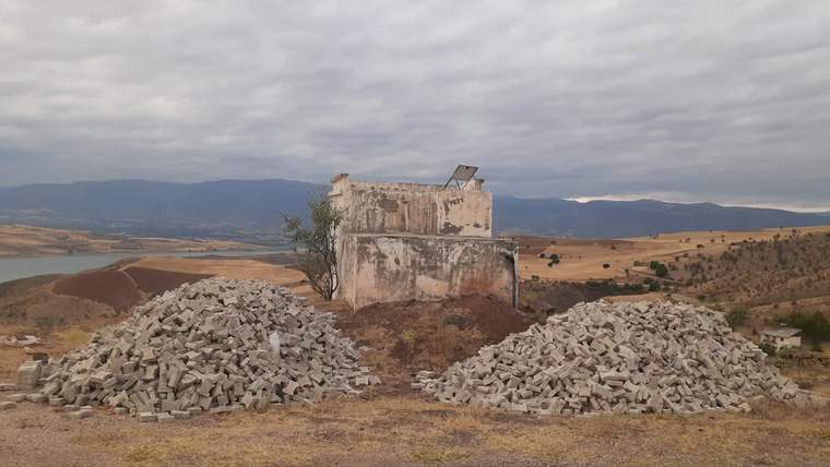 Kırsal arazide, eski bir taş yapının önünde iki büyük yığın halinde kilit parke taşları bulunuyor.