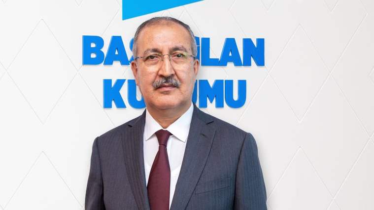 Koyu gri takım elbise giymiş, kravat takan bir kişi, arkasında "BASINLAN KURUMU" yazılı bir pano ile poz veriyor.