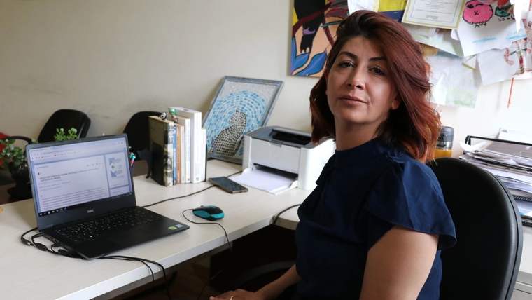 Çalışma masasında oturan bir kadın, açık dizüstü bilgisayar ve ofis eşyalarıyla kameraya bakıyor.