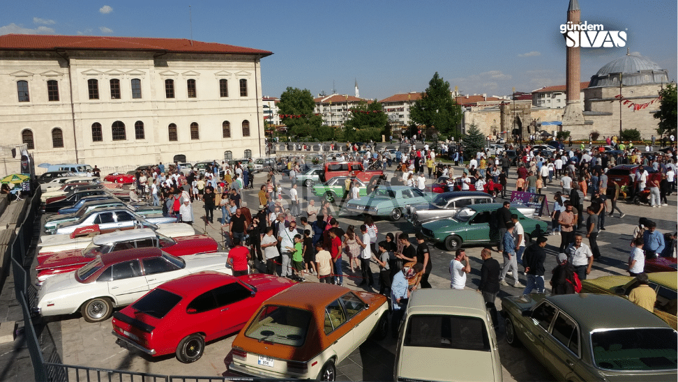 Çeşitli klasik otomobillerin arasında dolaşan kalabalık, tarihi taş binanın önündeki alanda görülüyor.