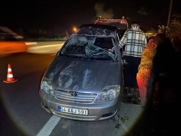 Kaza geçiren gri bir otomobil, yol kenarında duruyor; araçta hasar var ve yanındaki kişi inceleme yapıyor.