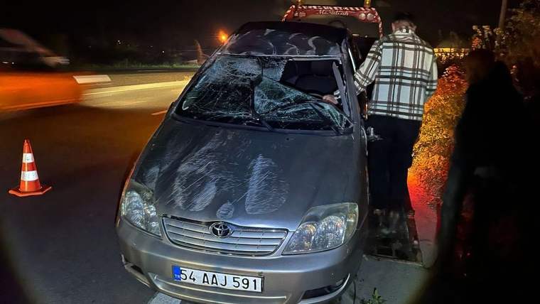 Kaza geçiren gri bir otomobil, yol kenarında duruyor; araçta hasar var ve yanındaki kişi inceleme yapıyor.
