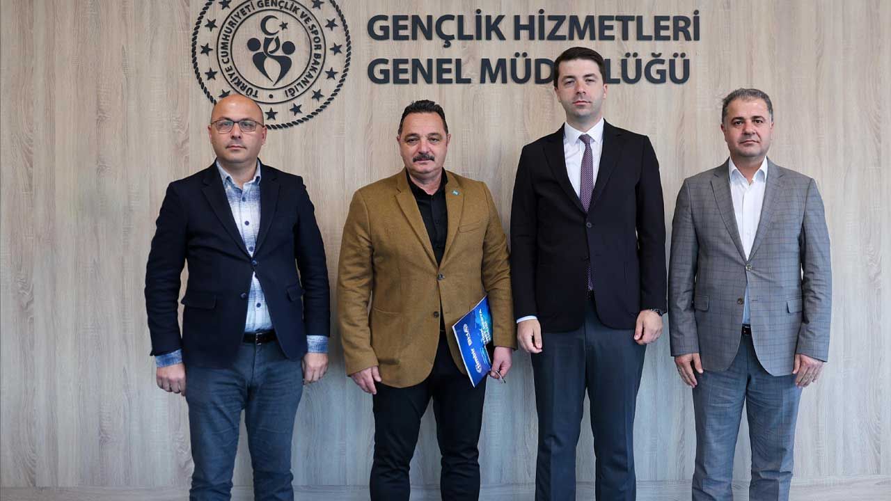 Gençlik Hizmetleri Genel Müdürlüğü yazısı önünde, dört erkek yan yana duruyor; birinin elinde mavi dosya var.