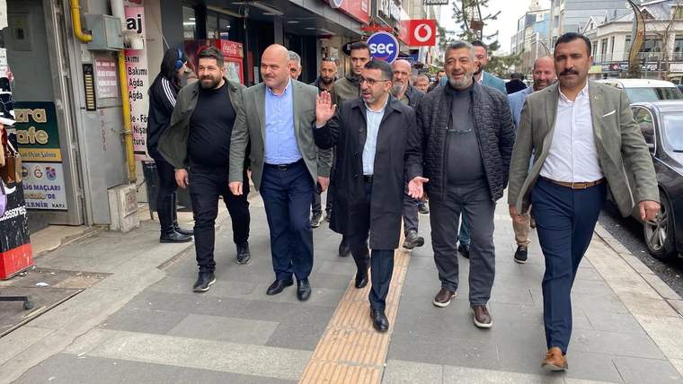 Bir grup erkek, dükkânların bulunduğu kaldırımda yürürken içlerinden biri el sallıyor.