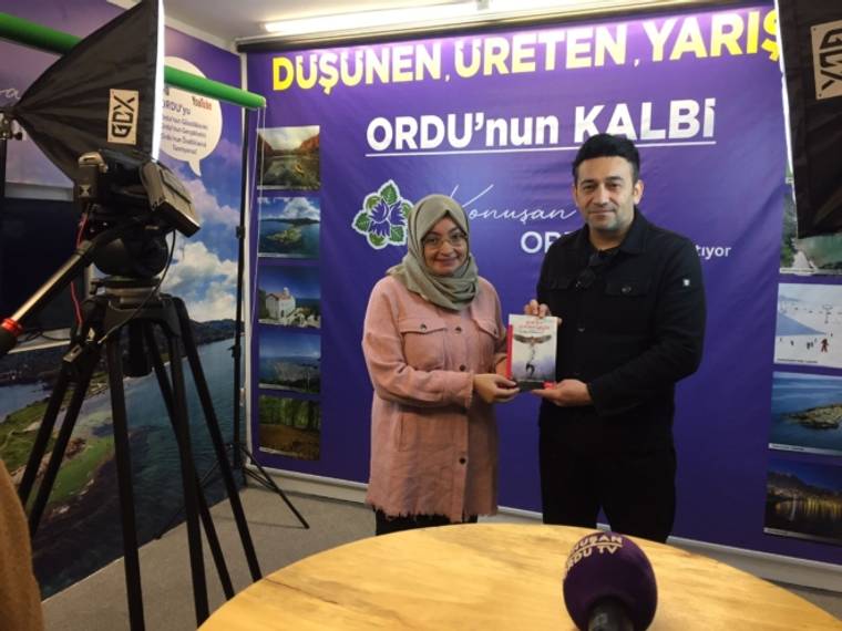 Bir kadın ve bir erkek, mor tanıtım panosu önünde birlikte kitap tutuyor; önde kamera ve mikrofon var.