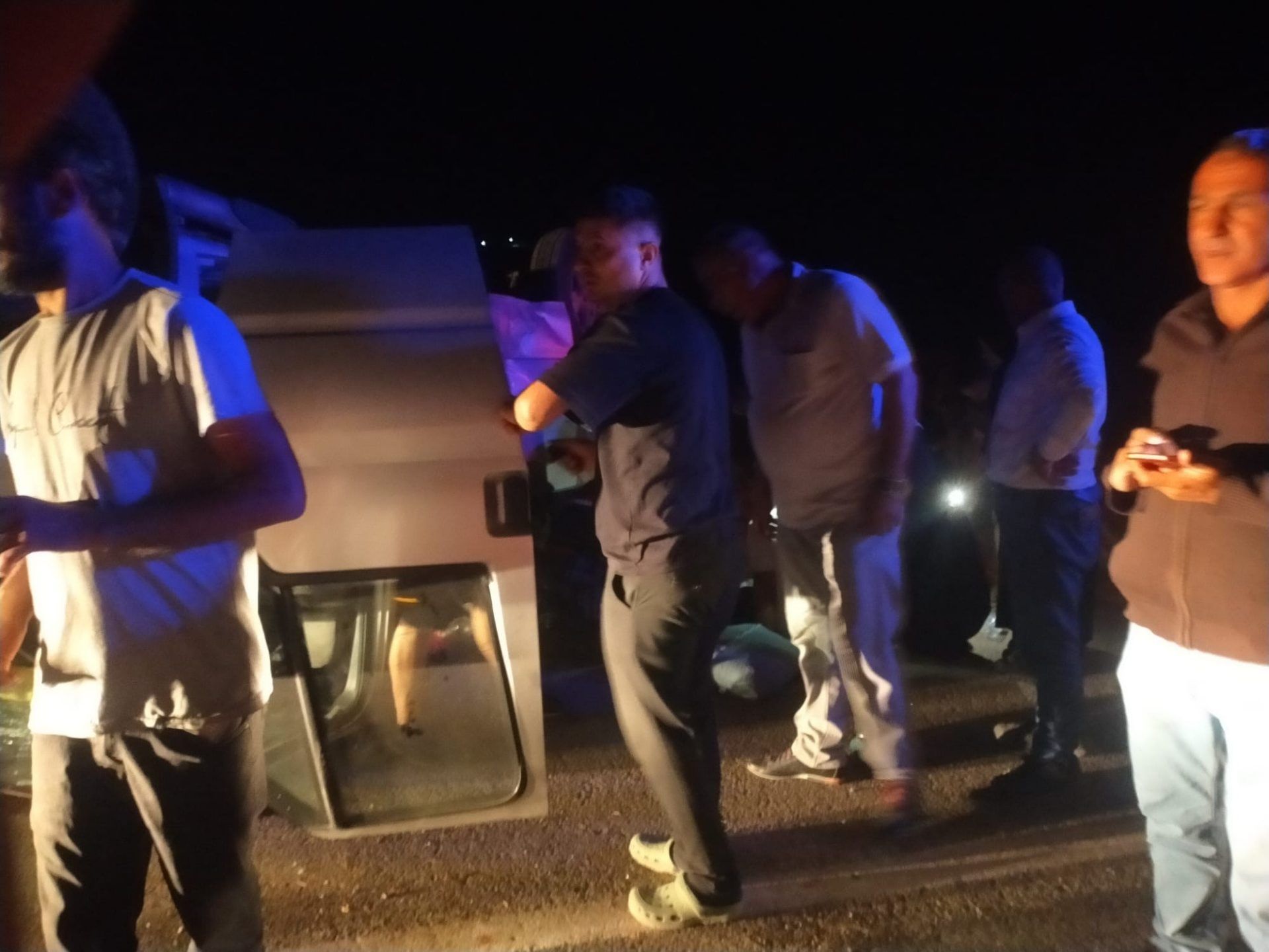 Gece karanlığında, yan yatmış beyaz bir aracın çevresinde duran birkaç kişi görülüyor.