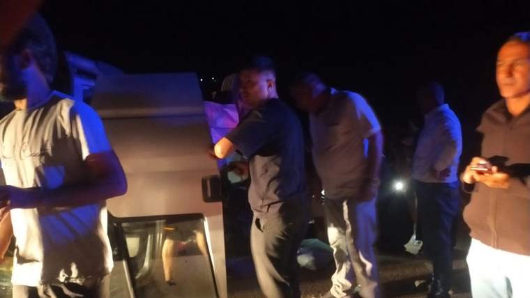 Gece karanlığında, yan yatmış beyaz bir aracın çevresinde duran birkaç kişi görülüyor.