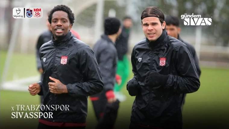 Siyah antrenman kıyafetleri giyen iki futbolcu sahada koşarken, arkalarında başka oyuncular görülüyor.