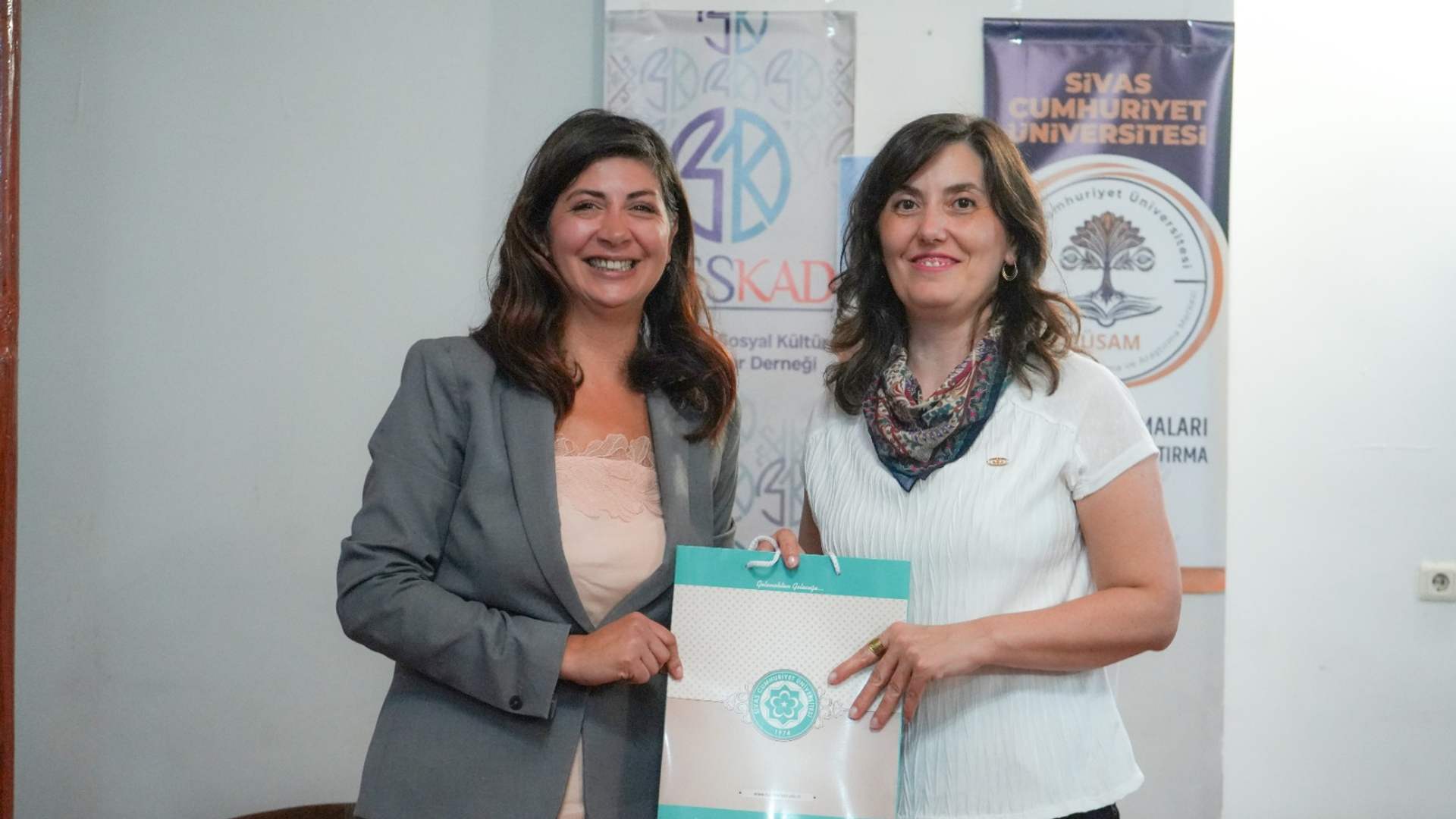 İki kadın, üzerinde belge bulunan turkuaz bir dosyayı tutarak kamera karşısında duruyor; arkada üniversite afişleri yer alıyor.