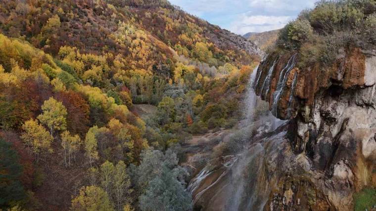 Sonbahar renklerine bürünen vadide, kayalık yamaçtan aşağı akan şelale ve çevresindeki ağaçlar görülüyor.