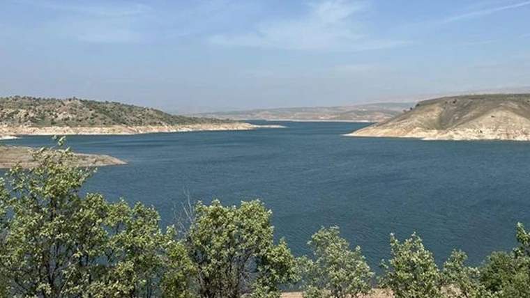 Kıyıları kuru toprakla çevrili geniş bir baraj gölü ve ön planda yeşil ağaçlar görülüyor.