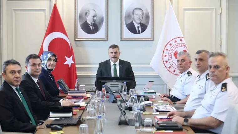 Toplantı masasında oturan bir grup kişi, Türk bayrağı ve amblemli beyaz bayrak önünde bir arada görülüyor.