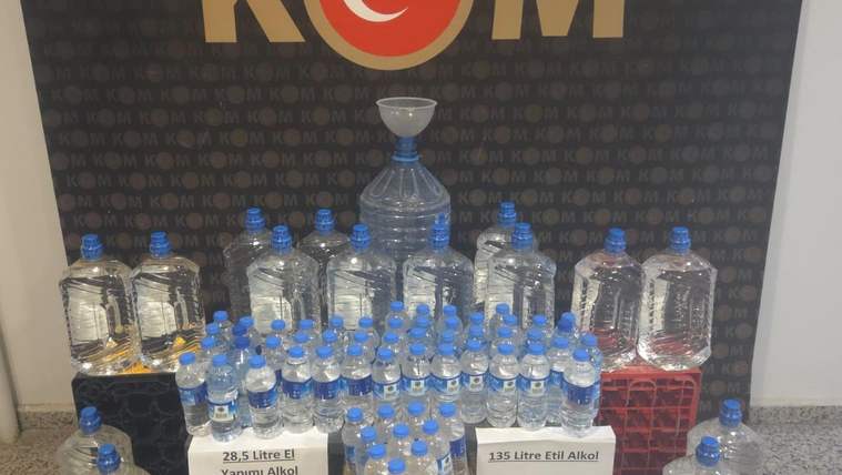 KOM yazılı pano önünde, çok sayıda şişe ve bidon ile üzerinde litre bilgileri bulunan alkol etiketleri görülüyor.
