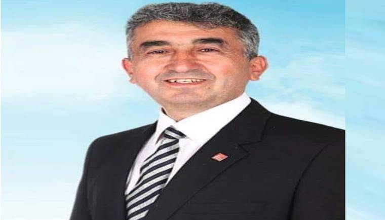 Takım elbiseli, kravatlı ve yakasında rozet bulunan bir erkek, açık mavi fon önünde gülümsüyor.