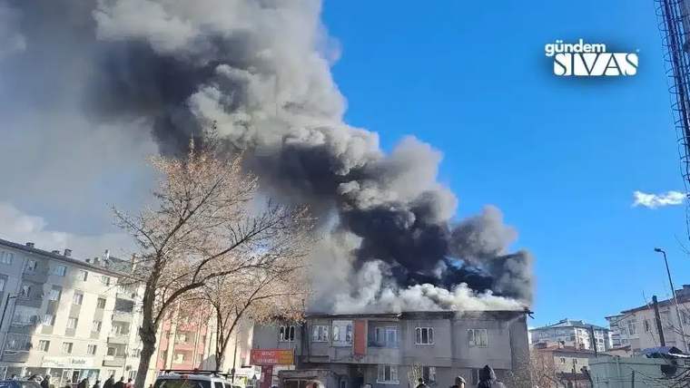 Bir binanın çatısından yoğun siyah duman yükseliyor, çevresinde ağaçlar ve araçlar var.