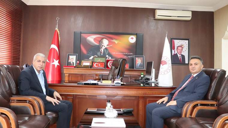 İki kişi, bir ofiste yan yana oturmuş, masanın arkasında Türk bayrağı ve Atatürk'ün resmi var.