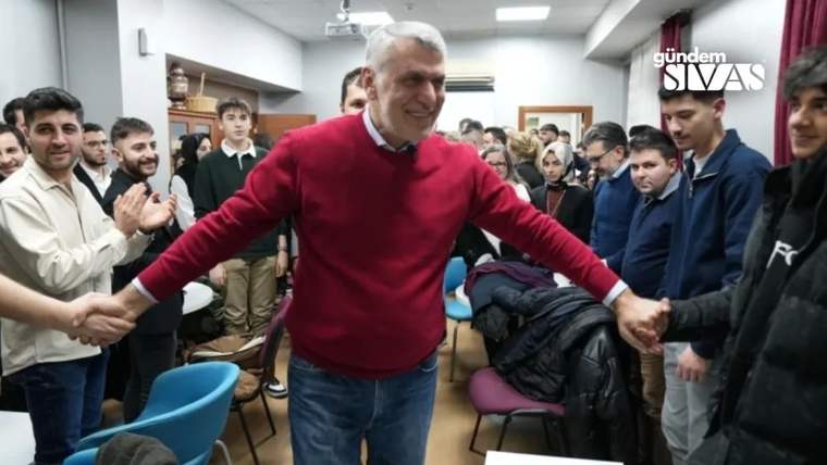 Kırmızı kazak giymiş bir kişi, elini havaya kaldırarak kalabalığın ortasında gülümsemekte.