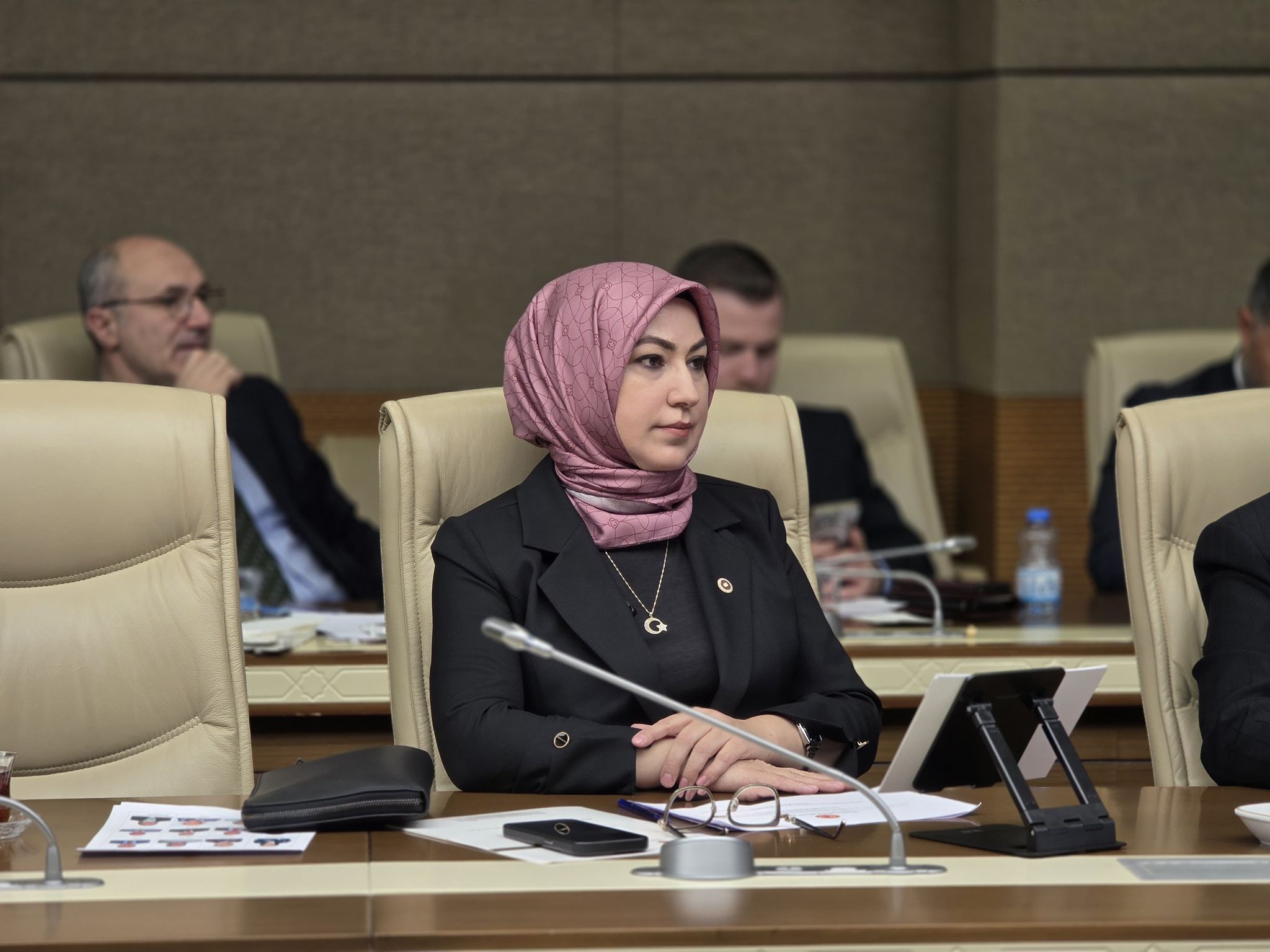 Başörtülü bir kadın, toplantı salonunda mikrofon ve belgelerin bulunduğu masada oturuyor.