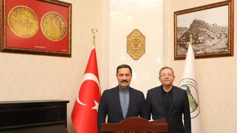 İki erkek kürsünün arkasında duruyor; yanlarında Türk bayrağı, başka bir bayrak ve siyah piyano bulunuyor.