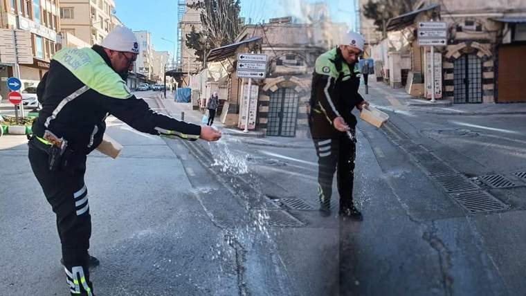 İki trafik görevlisi, buzla kaplı yola beyaz granüller serperken görülüyor; çevrede binalar ve tabelalar bulunuyor.