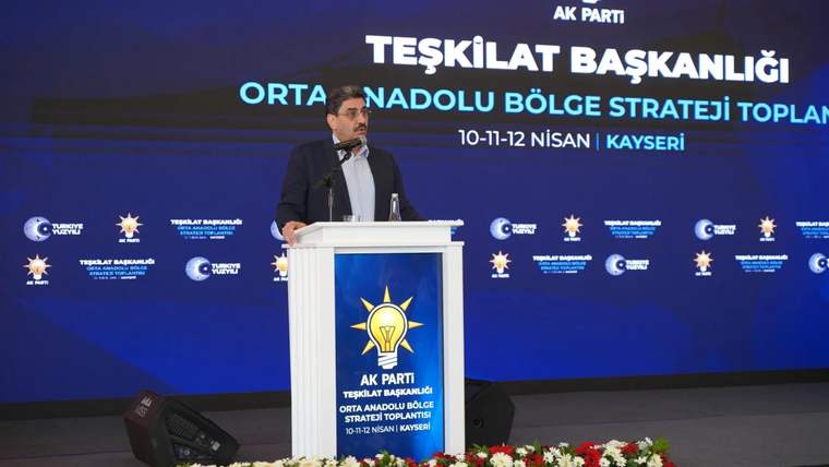 Mikrofonun önünde bir kişi, arkasında AK Parti'nin strateji toplantısı için hazırlanmış sahne dekoru var.