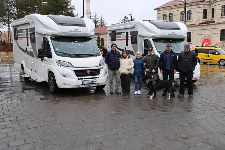 Yağmurla ıslanmış meydanda iki karavan, yanlarında duran altı kişi ve bir köpek görülüyor.