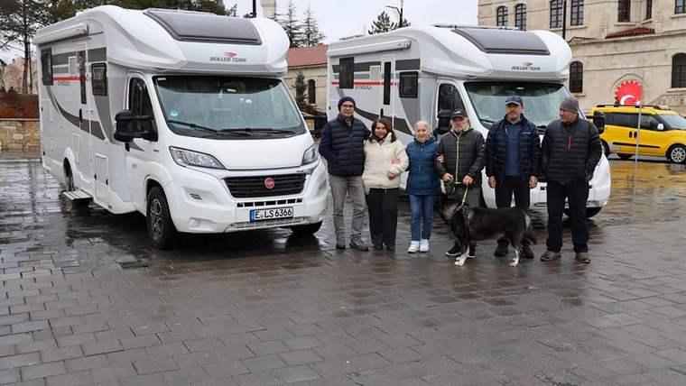 Yağmurla ıslanmış meydanda iki karavan, yanlarında duran altı kişi ve bir köpek görülüyor.