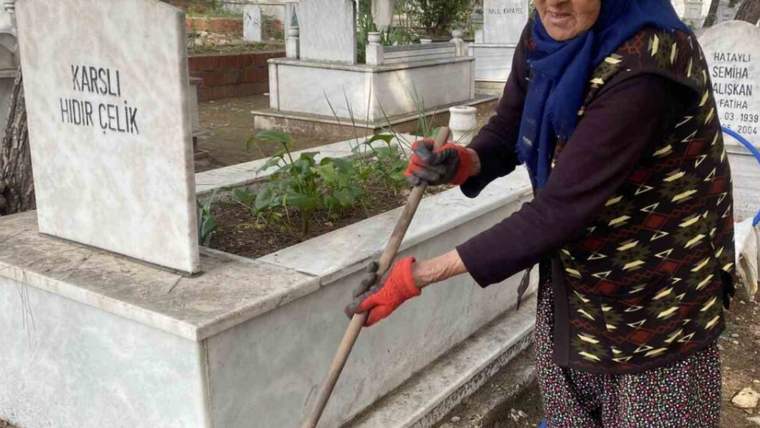 Başörtülü ve eldivenli bir kadın, mezarlıkta mezarların arasında uzun saplı bir aletle temizlik yapıyor.