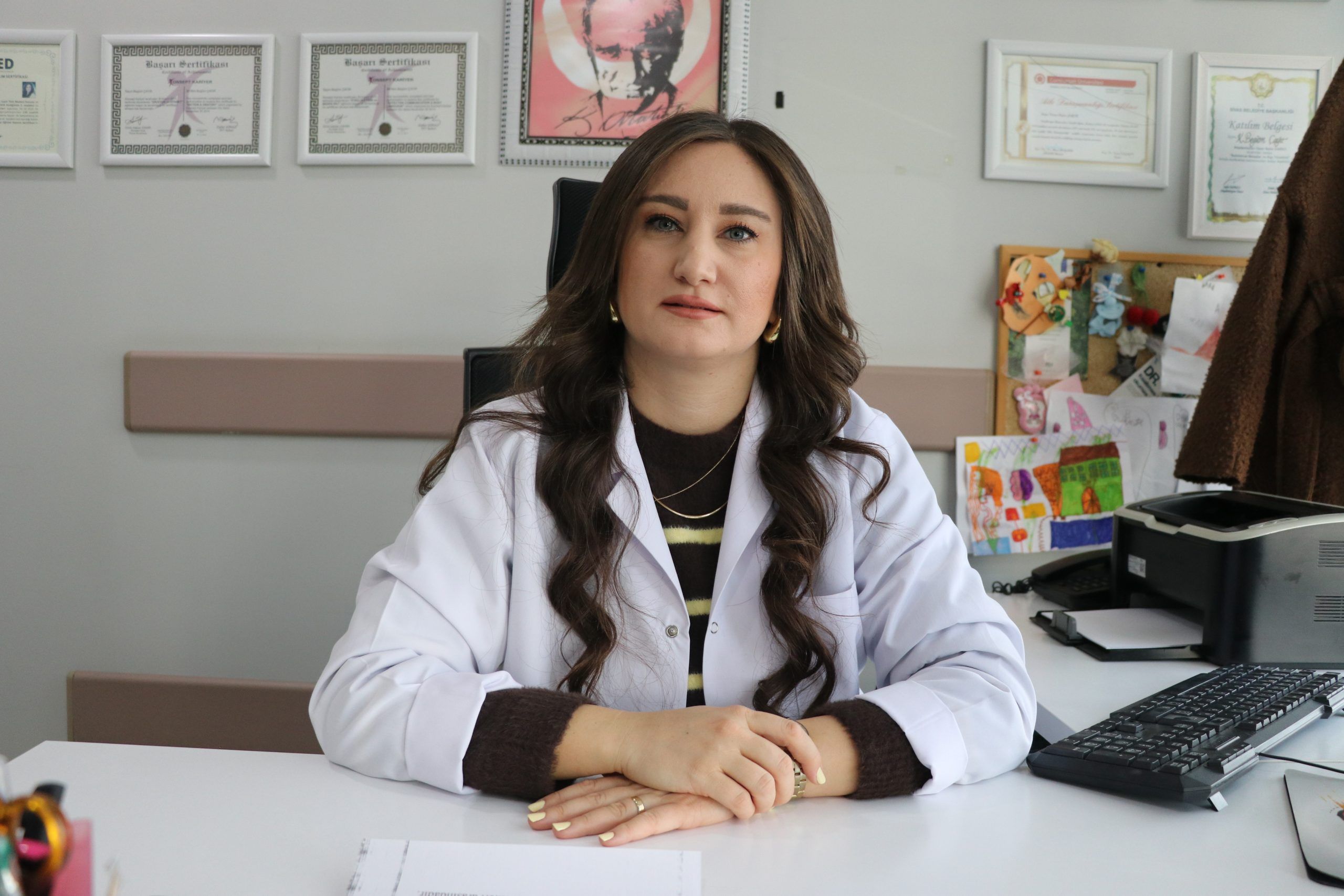 Beyaz önlük giymiş bir kadın, masa başında oturuyor ve kameraya bakıyor.