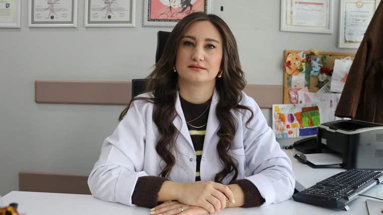 Beyaz önlük giymiş bir kadın, masa başında oturuyor ve kameraya bakıyor.